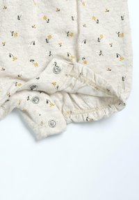 Combinaison en coton beige avec de petits motifs floraux jaunes, blancs et verts. Présente des fermetures à pression au bas et des ouvertures de jambe élastiquées.