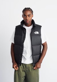Black puffer vest with a high collar and zip closure, featuring a white logo on the left chest, worn over a white t-shirt.
