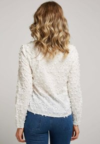 Blouse blanche à manches longues brodée, ornée de motifs floraux, d'accents texturés et avec un ourlet arrondi, associée à un jean en denim bleu.