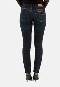 Vaqueros ajustados en denim de lavado oscuro con un desgaste sutil. Presentan bolsillos traseros, bolsillo con cremallera y parche de cuero marrón en la cintura.