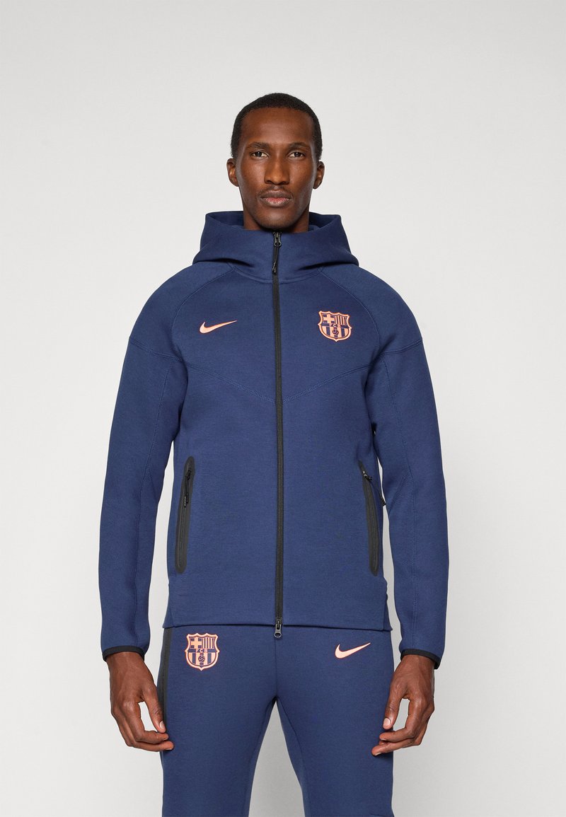 Veste à capuche marine avec des zippers noirs, des accents de logo en rose clair et un design sportif. Le matériau semble doux avec une texture lisse.