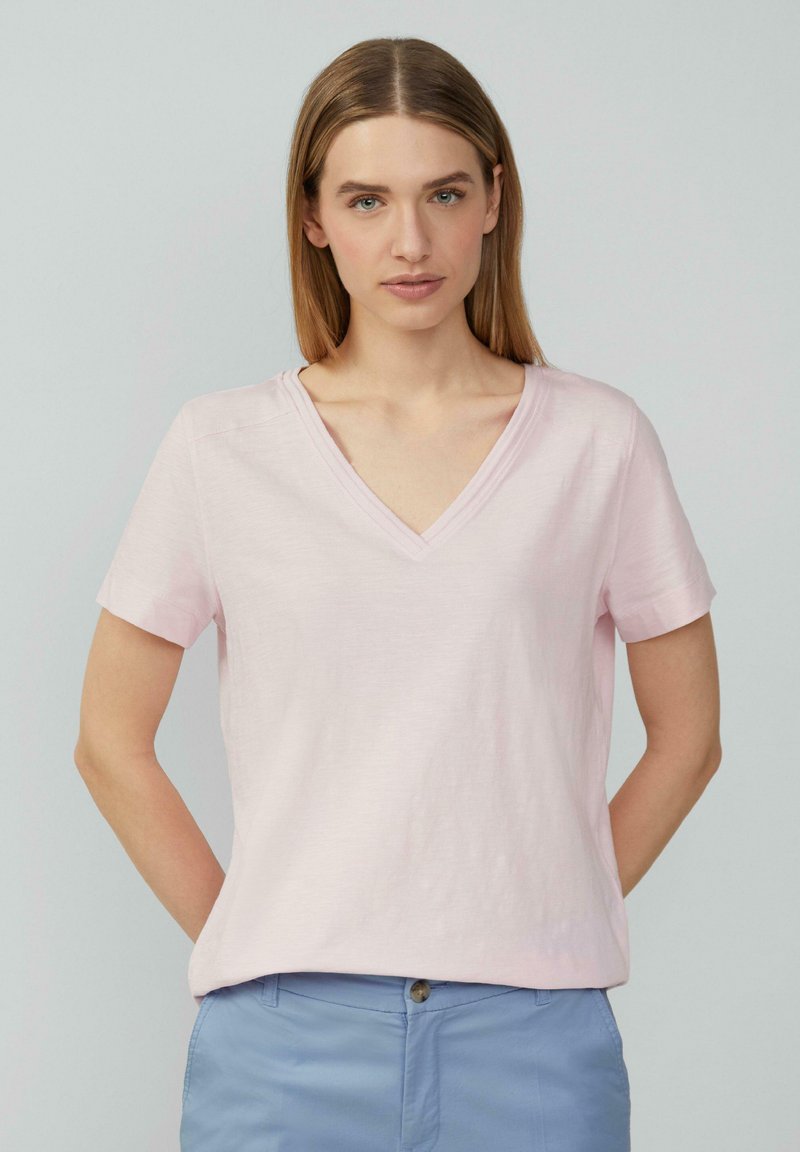 Lichtroze T-shirt met korte mouwen en V-hals, gemaakt van een zachte stof, met een ontspannen pasvorm en subtiele textuur. Gedragen met lichtblauwe broek.