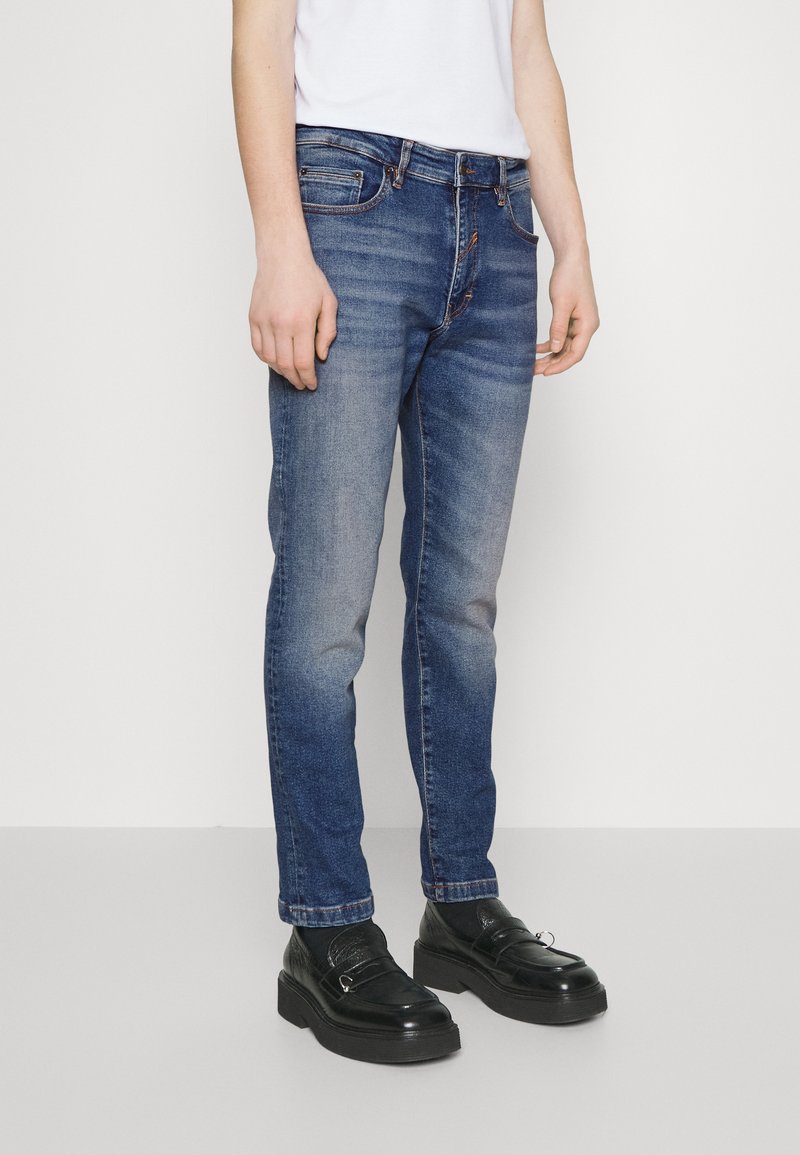 Jean en denim bleu avec un devant décoloré, coupe droite, cinq poches et des détails de couture subtils, associés à des mocassins noirs.