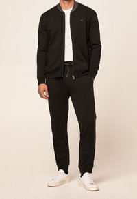 Chaqueta bomber negra con cremallera y puños ribeteados, combinada con pantalones jogger negros a juego y zapatillas blancas. Diseño simple y textura suave.