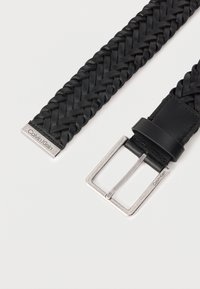 Πλεγτή μαύρη δερμάτινη ζώνη με ασημένια μεταλλική αγκράφα και διακριτική λεπτομέρεια με λογότυπο. Διαθέτει κομψή, επίπεδη υφή και μοντέρνο σχεδιασμό.