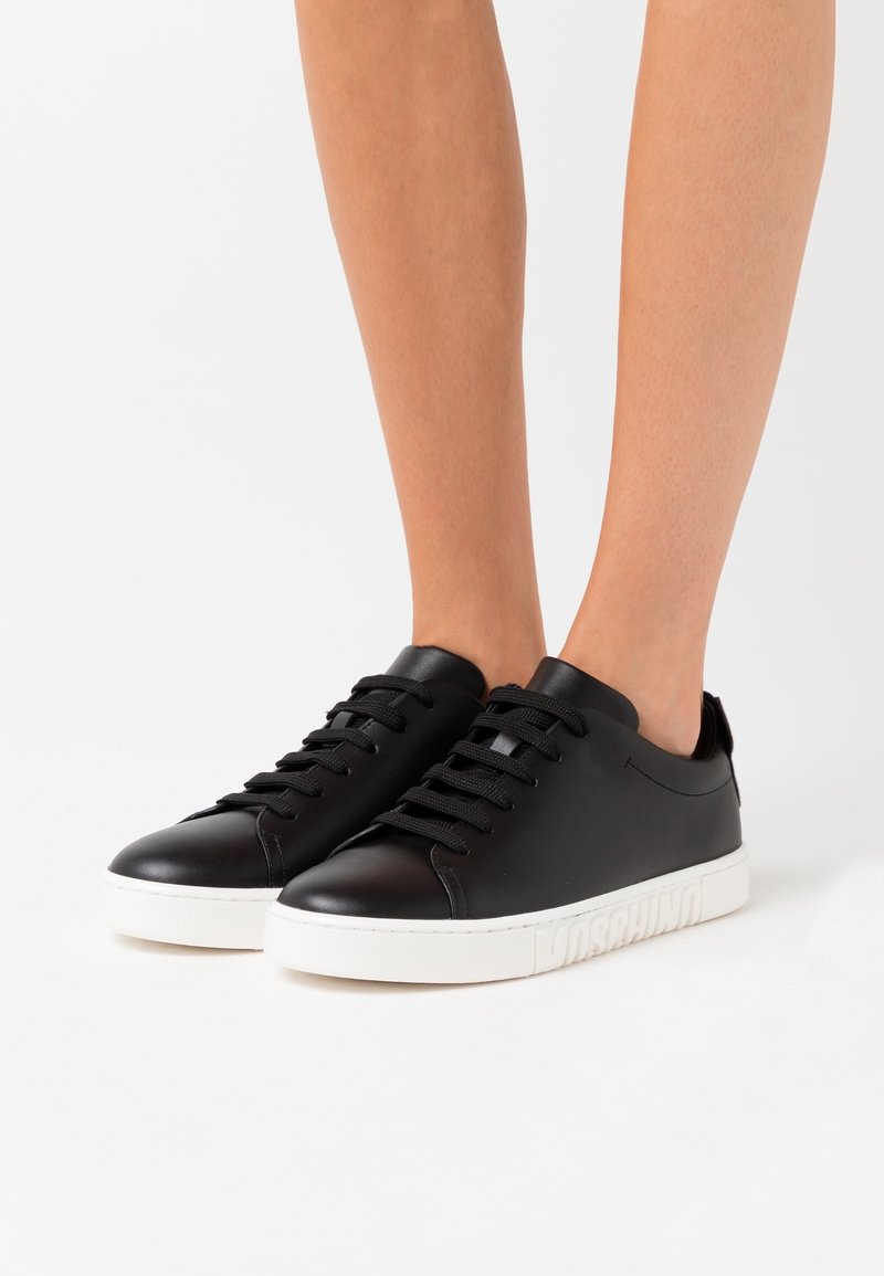 Jambes portant des baskets noires basses avec des semelles épaisses blanches et des lacets, debout sur une surface blanche.