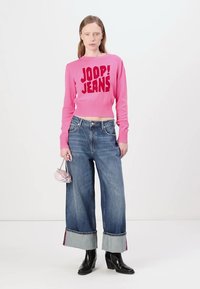 Suéter de punto corto en color rosa con la inscripción "JOOP! JEANS", acompañado de unos vaqueros de tiro alto de pierna ancha en denim azul con dobladillo en contraste, y botas negras de tobillo.