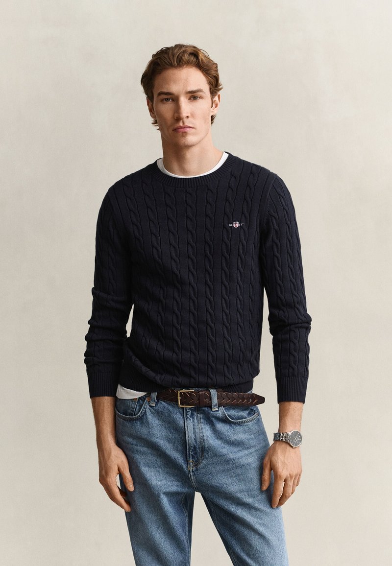 Young man wearing a navy blue cable knit sweater, light blue jeans, brown braided belt, and a silver wristwatch, standing against a neutral background.