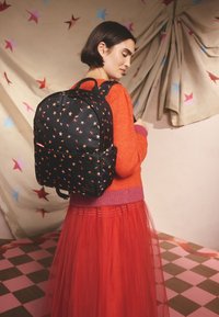 Moteris oranžiniais megztiniu ir raudona suknele, nešiojanti juodą kuprinę su žvaigždžių raštu, stovinti priešais smėlio spalvos sieną su žvaigždžių dekoracijomis.