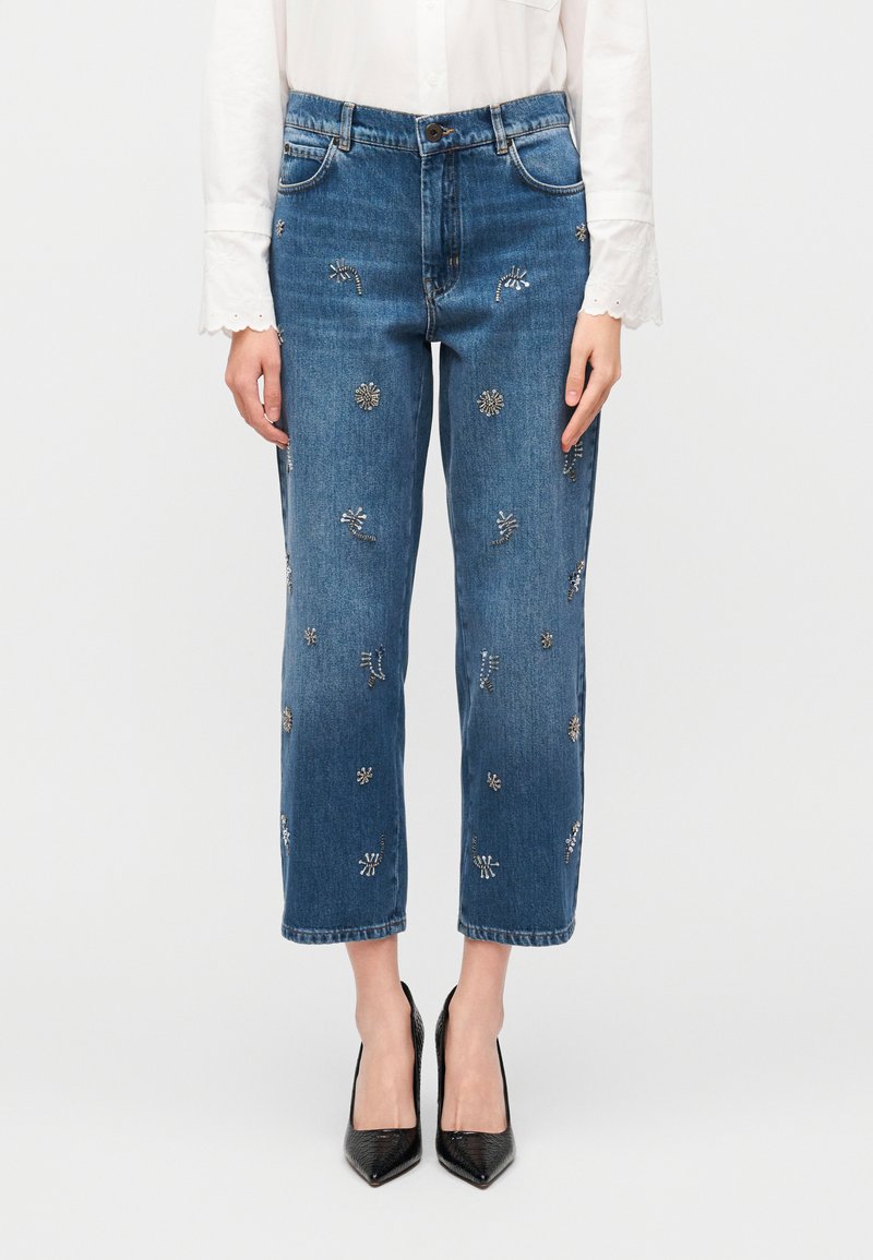 Μπλε τζιν με χαλαρή εφαρμογή, διακοσμημένο με ασημένια κεντήματα λουλουδιών και κοντό μήκος. Συνδυάζεται με μαύρες γόβες με μυτερή μύτη.