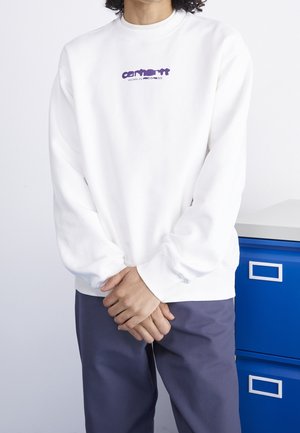 Person iført hvid sweatshirt med lilla "Carhartt Work in Progress" logo og mørke bukser, stående med hænderne foldet foran.