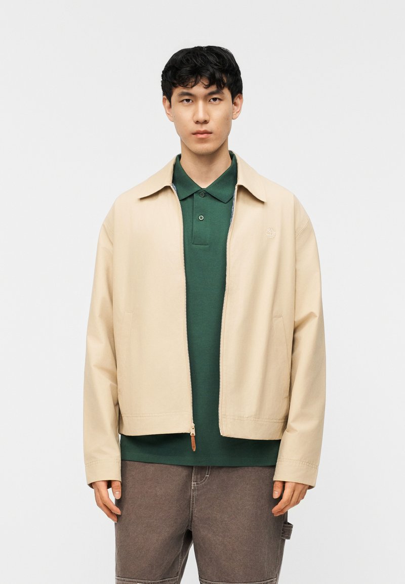 Νέος άνδρας που φοράει μπεζ μπουφάν με φερμουάρ πάνω από πράσινο πόλο πουκάμισο και καφέ παντελόνια, στέκεται μπροστά σε απλό λευκό φόντο.