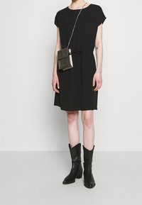 Persona vistiendo un vestido negro de manga corta, botas negras hasta la mitad de la pantorrilla y un pequeño bolso cruzado metálico con una cadena.