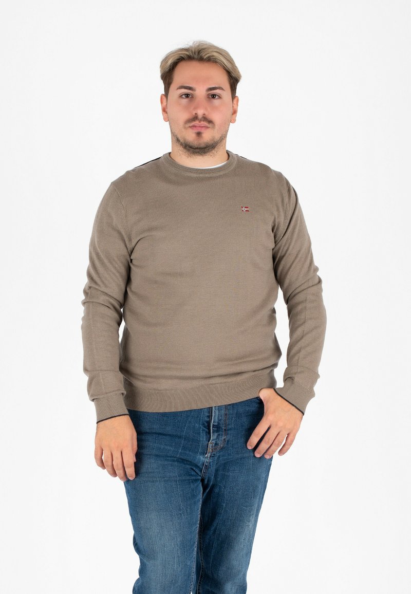 Uomo con capelli corti chiari che indossa un maglione marrone e jeans blu, in piedi con una mano in tasca su uno sfondo bianco.