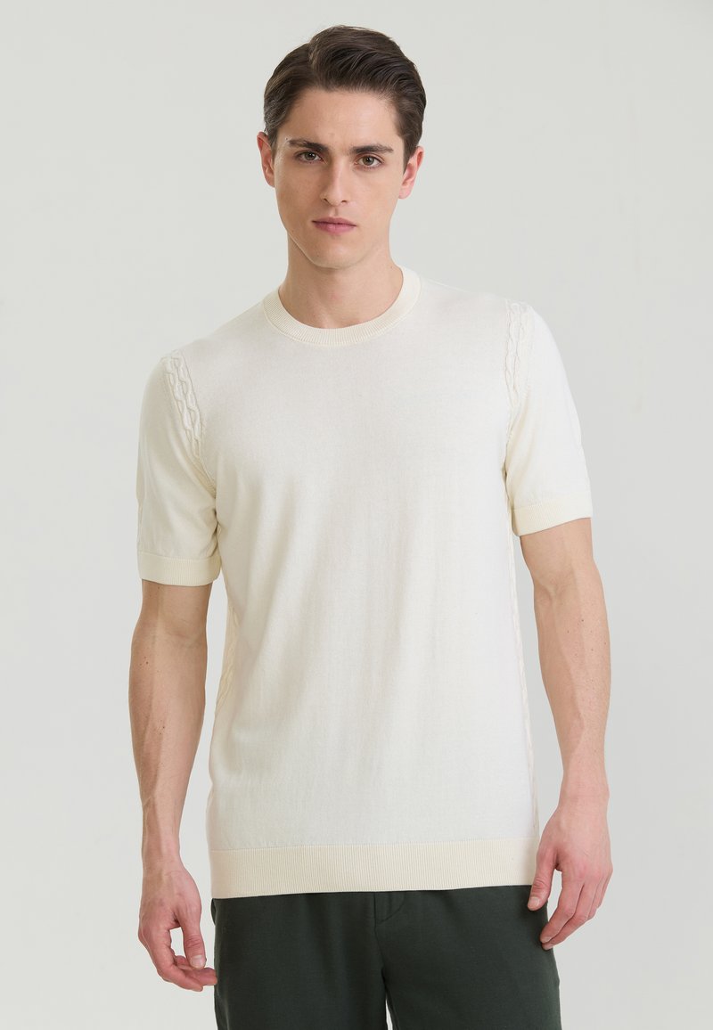 Jeune homme aux cheveux courts et foncés, portant un pull en maille crème à manches courtes et un pantalon foncé, debout devant un fond clair uni.
