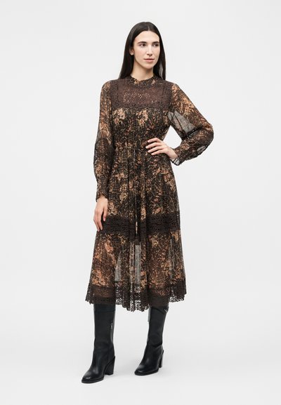 Γυναίκα στέκεται φορώντας μακρυμάνικο, καφέ φόρεμα με λουλουδένιο σχέδιο μέχρι τη μέση των ποδιών, με μαύρες δαντελένιες λεπτομέρειες και μαύρες, ψηλές, ψηλοτάκουνες μπότες μέχρι το γόνατο.