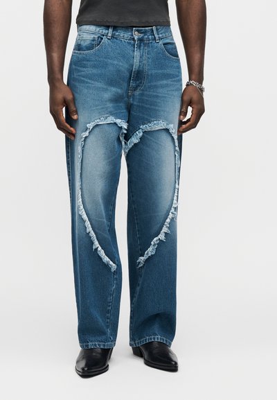 Muž v modrých džínsoch s veľkými vyfrkanými výstrihmi v tvare srdca na stehnách, čiernej košeli, čiernych topánkach a striebornej náramkovej retiazke.