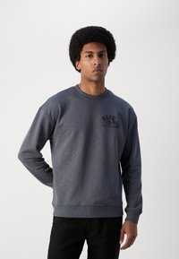 Sivi dukserica s okruglim izrezom i rebrastim rukavima. Ima crni grafički dizajn na prsima. Mekana tekstura s ležernim krojem.