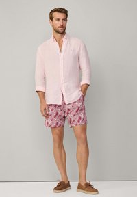 Camicia button-up di lino rosa chiaro, pantaloni corti rossi con motivi di foglie di palma, scarpe slip-on marroni, in piedi contro uno sfondo neutro.
