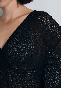 Abito in pizzo nero con dettagli intricati a taglio ovale, design con profondo scollo a V e cintura arricciata. Tessuto leggero, con strato sottostante inibito.