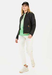 Black, quilted jacket with a front zipper, paired with a green sweatshirt and white pants, complemented by light sneakers and a black cap.