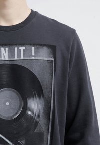 Felpa grigia scura con una grafica stampata di un disco in vinile e il testo "PLAY IT!" in bianco. Collo rotondo, texture morbida, vestibilità casual.