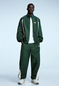 Vihreä verryttelytakki, jossa on violetteja ja kermaisia yksityiskohtia, yhdistettynä samaa vihreää sävyä oleviin housuihin. Molemmat vaatekappaleet ovat väljästi istuvia ja valmistettu kevyestä materiaalista.