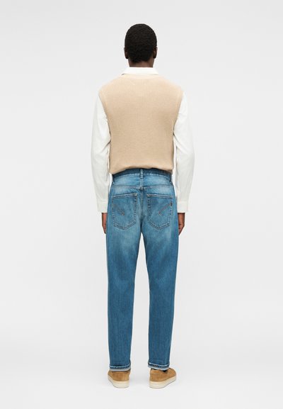 Άνδρας που φοράει μπεζ πλεκτό γιλέκο πάνω από λευκό πουκάμισο και μπλε τζιν, στέκεται γυρισμένος με την πλάτη σε απλό φωτεινό φόντο.