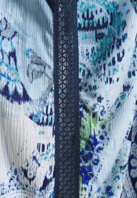 Tissu bleu clair avec des motifs abstraits bleus, verts et noirs, comprenant une bordure verticale en dentelle bleu marine à motif géométrique.