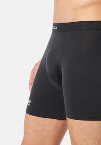 Zbliżenie na osobę noszącą czarne dopasowane bokserki, ukazujące uda do połowy i talia z małym logo na lewej nodze.