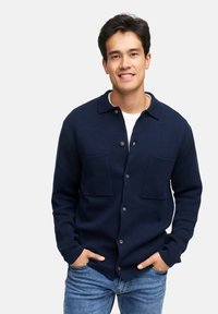 Navy gestrickte Strickjacke mit Knopfleiste, zwei Brusttaschen und Kentkragen; über einem weißen Hemd getragen, kombiniert mit blauen Jeans.