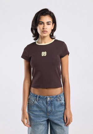 Young woman wearing a dark brown crop top with cream trim and number 89 print, paired with blue high-waisted jeans, standing against white background.