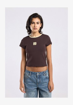 Young woman wearing a dark brown crop top with cream trim and number 89 print, paired with blue high-waisted jeans, standing against white background.