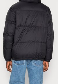 Giubbotto pesante nero con design a trapunta, cappuccio e polsini a coste. Tessuto lucido e liscio con vestibilità comoda, abbinato a jeans azzurri chiari.