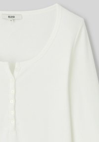 Haut à manches longues blanc à cannelures, avec une texture douce, présentant un col rond et une patte de boutonnage à l'avant. Étiquette de la marque visible.