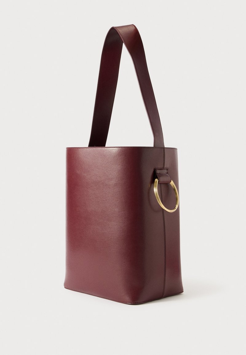 Sac en cuir bordeaux avec une forme structurée, une large bandoulière et un accent en anneau doré sur le côté. Texture lisse avec un minimum de détails.