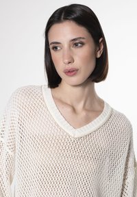 Cremefarbener Strickpullover mit einem offenen Netzdesign, V-Ausschnitt und gerippten Kanten. Strukturierter Stoff mit einem leichten und luftigen Aussehen.