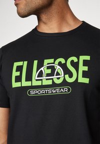Černé tričko s krátkým rukávem obsahující velký zelený nápis "ELLESSE" a "SPORTSWEAR", s bílým logem. Měkká, hladká textura.