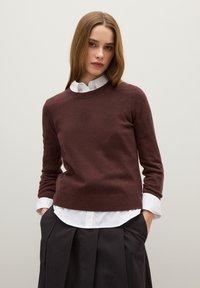 Maglione in maglia marrone con maniche lunghe e collo rotondo, indossato sopra una camicia bianca con colletto, abbinato a una gonna nera plissettata.