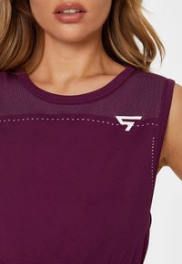 Gros plan d'une femme portant un haut de sport violet sans manches avec un empiècement en mesh et un logo blanc sur la poitrine.