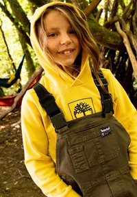 Un sweat à capuche jaune avec un logo d'arbre noir, associé à des salopettes vert olive avec des bretelles réglables et une poche avant. Fond en milieu naturel.