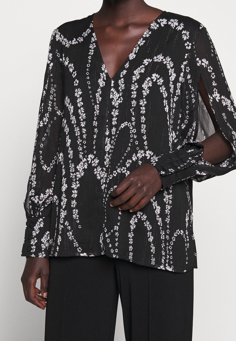 Blusa nera con motivo floreale bianco, scollatura a V, maniche lunghe e polsini con bottone. Tessuto trasparente con finitura testurizzata.