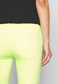 Jeans amarillos neón con un ajuste ajustado, bolsillos traseros y detalles de costura. El material parece elástico y tiene una textura suave.