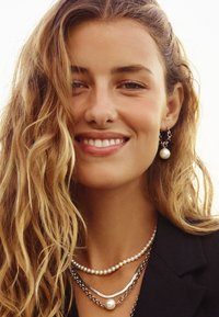 Silver- och pärlsmycken på en modell, med lager-på-lager halsband med kedjor och en enskild pärla som accenter, mot en ljus bakgrund.