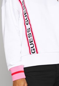 Sudadera blanca con el logo "GUESS" en negro y rosa en el lateral, con puños acanalados rosas y un corte holgado.