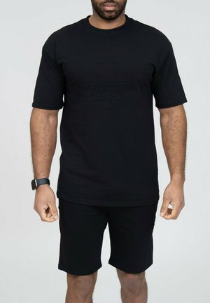 Homme portant un t-shirt noir à manches courtes avec le logo embossé "#ICON ORIGINAL" et un short noir assorti, debout devant un fond uni.