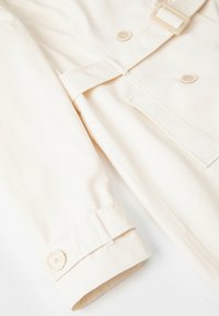 Trench coat beige chiaro con vita regolabile, chiusure a bottoni e dettagli ai polsini con bottoni. Tessuto liscio e design su misura.