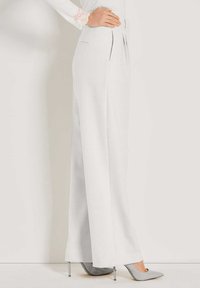 Vrouw in hoge taille, wijde witte broek en grijze, puntige hoge hakken, met de hand op de heup, tegen een effen achtergrond.