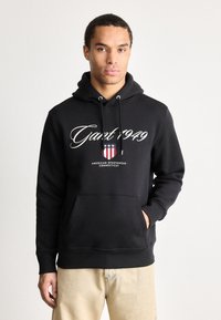 Tânăr îmbrăcat cu un hanorac negru cu inscripția "Gant 1949" și un logo de îmbrăcăminte sportivă americană, purtând pantaloni bej, stând în fața unui fundal simplu.