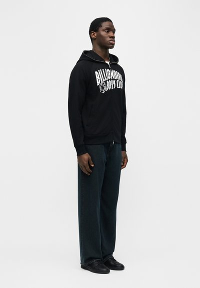 Άνδρας που φοράει μαύρο φούτερ με φερμουάρ και τη φράση «Billionaire Boys Club», σκούρα τζιν με φαρδιά μπατζάκια και μαύρα παπούτσια, στέκεται μπροστά σε λευκό φόντο.